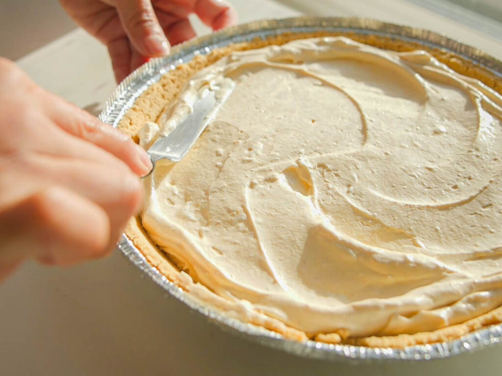 A person spreads creamy filling over a pie crust in a foil dish with a knife. The pie is on a wooden surface, and the sunlight casts a warm glow over the scene.